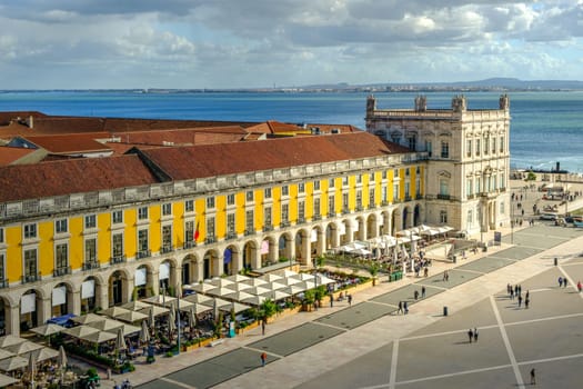 Scenic view of Commerce Square or Praca do Comercio in Lisbon, Portugal, iconic yellow Pombaline architecture, bustling outdoor cafes, Tagus River, city life, historic downtown, sunny day, sky with clouds. Sun ray over building, travel destination