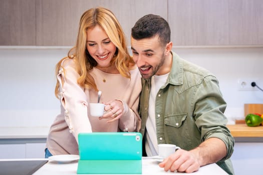 Smiling couple enjoying coffee together while using a tablet in their kitchen. They share a joyful moment, exploring content or making plans in a cozy and bright domestic environment.