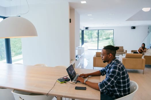African American influencer in glasses sitting at a table in a modern living room, using a laptop for business video chat, conversation with friends and entertainment