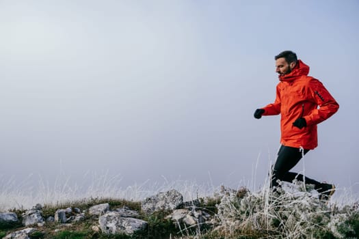 A man in athletic wear climbs a rocky hill in foggy weather. Dressed in a red jacket and black leggings, he is captured mid stride in a determined motion against a pale, misty background.