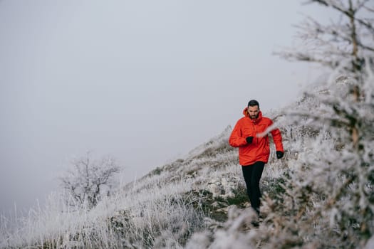 A man in athletic wear climbs a rocky hill in foggy weather. Dressed in a red jacket and black leggings, he is captured mid stride in a determined motion against a pale, misty background.