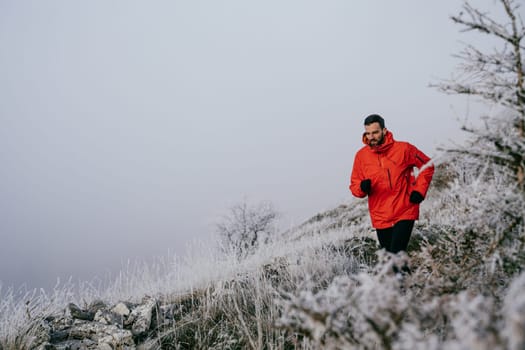 A man in athletic wear climbs a rocky hill in foggy weather. Dressed in a red jacket and black leggings, he is captured mid stride in a determined motion against a pale, misty background.