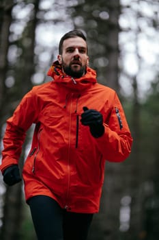 A man wearing a red jacket is jogging on a forest trail surrounded by tall pine trees and autumn foliage. The serene woodland path suggests a peaceful outdoor activity in nature during the fall season.