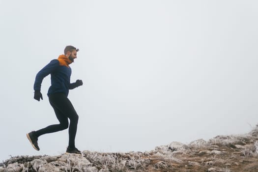 A man in athletic wear climbs a rocky hill in foggy weather. Dressed in a blue jacket and black leggings, he is captured mid-stride in a determined motion against a pale, misty background.