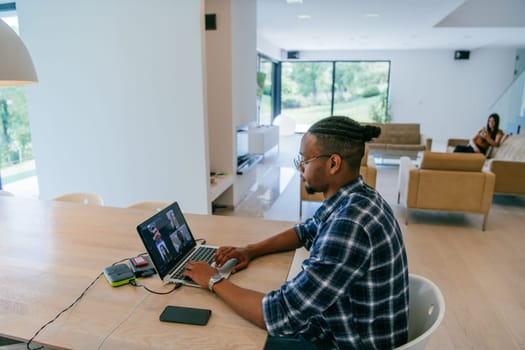 African American influencer in glasses sitting at a table in a modern living room, using a laptop for business video chat, conversation with friends and entertainment