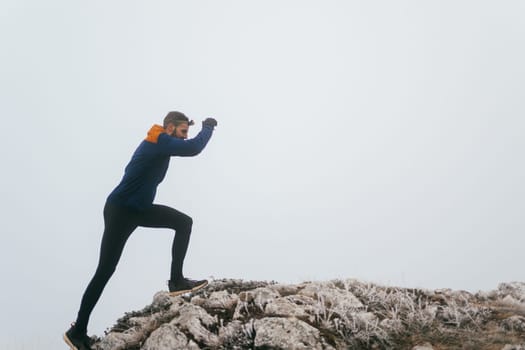 A man in athletic wear climbs a rocky hill in foggy weather. Dressed in a blue jacket and black leggings, he is captured mid-stride in a determined motion against a pale, misty background.