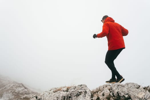 A man in athletic wear climbs a rocky hill in foggy weather. Dressed in a red jacket and black leggings, he is captured mid stride in a determined motion against a pale, misty background.