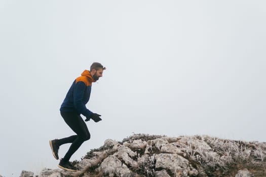 A man in athletic wear climbs a rocky hill in foggy weather. Dressed in a blue jacket and black leggings, he is captured mid-stride in a determined motion against a pale, misty background.
