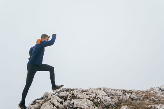 A man in athletic wear climbs a rocky hill in foggy weather. Dressed in a blue jacket and black leggings, he is captured mid-stride in a determined motion against a pale, misty background.