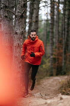 A man wearing a red jacket is jogging on a forest trail surrounded by tall pine trees and autumn foliage. The serene woodland path suggests a peaceful outdoor activity in nature during the fall season.