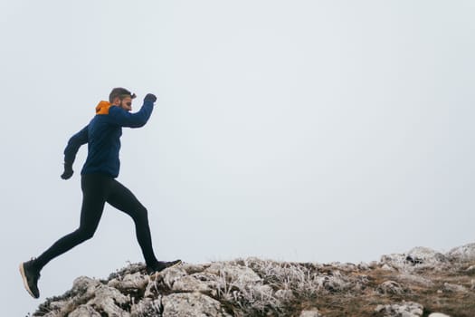 A man in athletic wear climbs a rocky hill in foggy weather. Dressed in a blue jacket and black leggings, he is captured mid-stride in a determined motion against a pale, misty background.