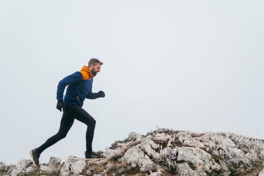 A man in athletic wear climbs a rocky hill in foggy weather. Dressed in a blue jacket and black leggings, he is captured mid-stride in a determined motion against a pale, misty background.