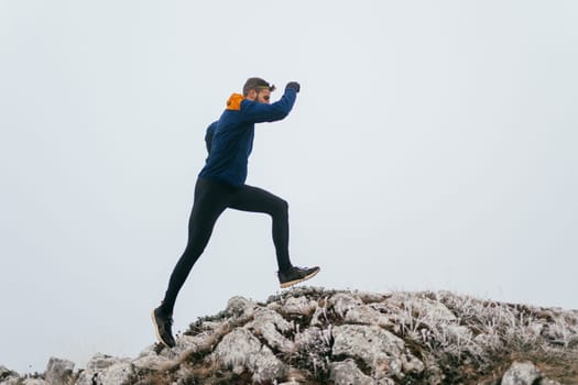 A man in athletic wear climbs a rocky hill in foggy weather. Dressed in a blue jacket and black leggings, he is captured mid-stride in a determined motion against a pale, misty background.