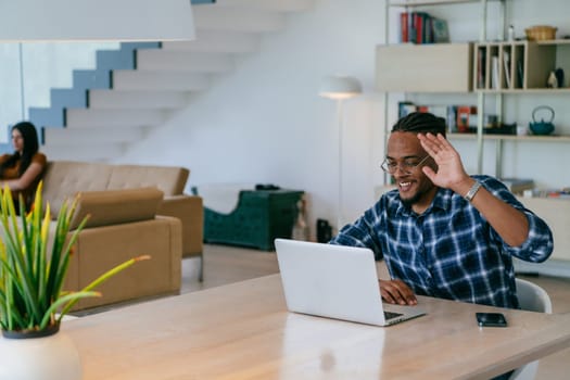African American influencer in glasses sitting at a table in a modern living room, using a laptop for business video chat, conversation with friends and entertainment