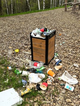 Overflowing trash bin in a park surrounded by scattered litter. Various items like cups, cans, and bags are on the ground, highlighting pollution and environmental neglect. Made with a smartphone.