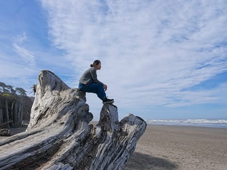 A middle aged woman sits on top of a large driftwood tree on the beach near Kalaloch area of Washington state.