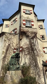 Innsbruck, Austria - March 28, 2025: Historic building with red and white window shutters painted in the colors of the Austrian flag, adorned with ivy and a bronze statue, highlighting architectural elegance and artistic expression