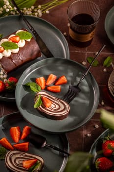 Chocolate swiss roll cake slices served on plates, garnished with fresh strawberries, mint leaves, and whipped cream, accompanied by a glass of coffee