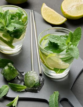 Glass of mojito cocktail with fresh mint leaves, lime slices, and ice cubes on a dark stone background. Refreshing summer drink with stainless steel straws