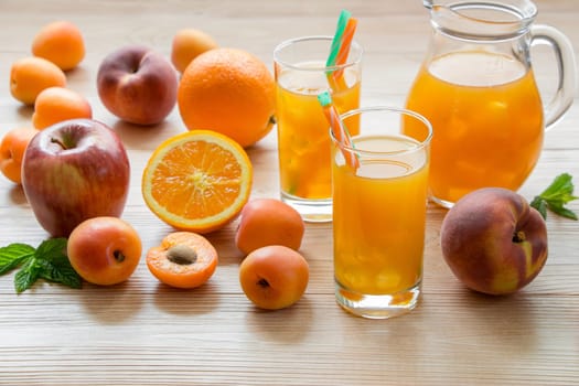 Fruit juice in glasses with ice near the jar of juice and scattered apricots, peaches, apple, orange on a light wooden background. Apricot peach apple orange juice with ice. Horizontal.