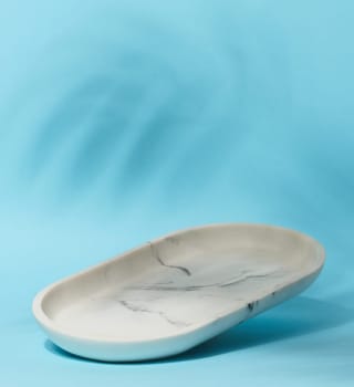 An empty, oval shaped soap dish made of white marble with gray veining sits on a light blue background with soft, blurred leaf shadows