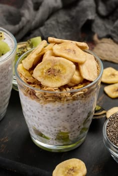 Two glasses of chia seed pudding, one topped with diced kiwi and the other with granola and banana chips, are on a dark surface with ingredients