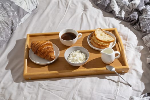 Delicious breakfast tray with croissant, bread, coffee, and cottage cheese set on a cozy bed in natural morning light.