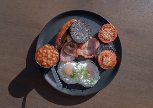 Top view of The full English Breakfast includes grilled bacon, sausage, baked beans, mushroom, tomatoes, black pudding is a sausage made with pork and flavored with pig blood and two hen's fried eggs with a runny yolk and watercress served on ceramic plate. use it as your Wallpaper, Poster and Space for text, Selective focus.