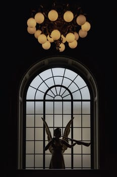 London, UK - May 07, 2025 - Silhouette view of Outstanding Angel of Gaiety statue (Spirit of Gaiety) in front of Large semi oval glass window and Hanging lantern from the ceiling on top at the Victoria and Albert Museum. Abstract interior design, use it as your Wallpaper, Poster and Space for text, Selective focus.