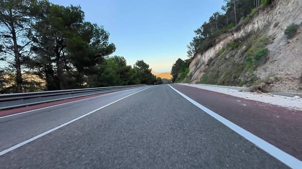 The vibrant sunset skies beautifully stretch over a winding road that is bordered by lush trees and rocky hills