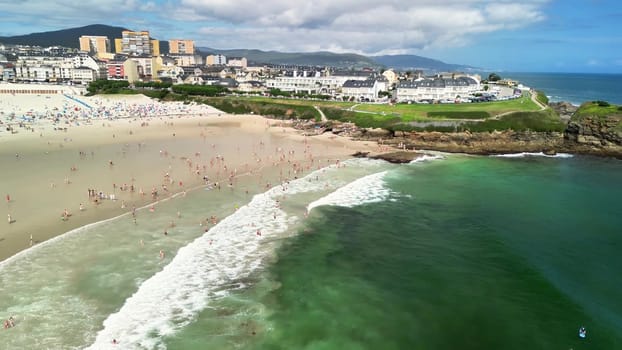 An aerial view shows a beautiful beach with gentle waves, clear water, and sunbathers under a blue sky