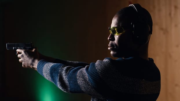 Person wearing safety goggles loading pistol bullets magazine during firearm practice session at shooting range. African american man reloading gun, learning self defense, pointing weapon, camera B