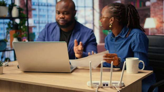 Black irritated man and woman rebooting their wireless router and checking network settings, feeling angry with the struggles to resolve an internet outage disrupting their tasks. Camera A.
