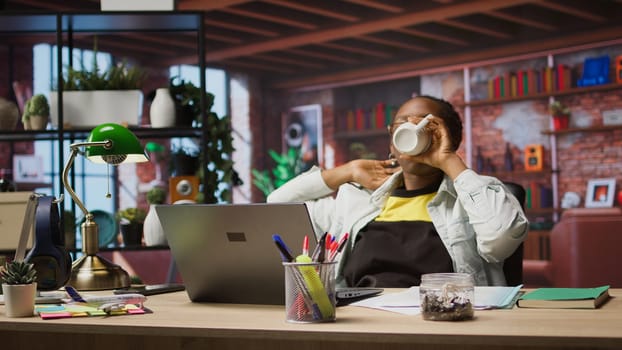 Happy woman drinking coffee, enjoying watching TV show during relaxation time at home. Cheerful person sitting at desk entertained by engrossing television series on laptop, camera A