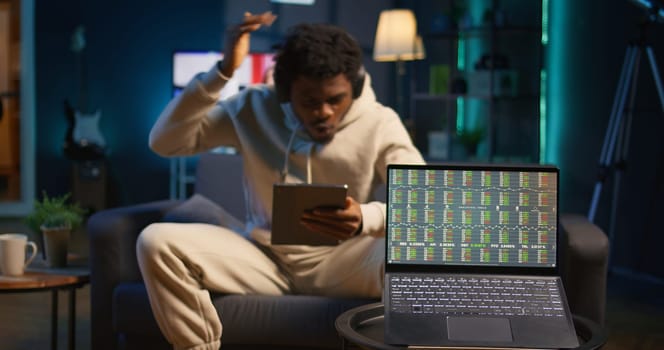 African american person using his tablet laying on the couch while he pays attention to laptop with stock market data trends. Focused man comparing trading graphs numbers before investing. Camera B.