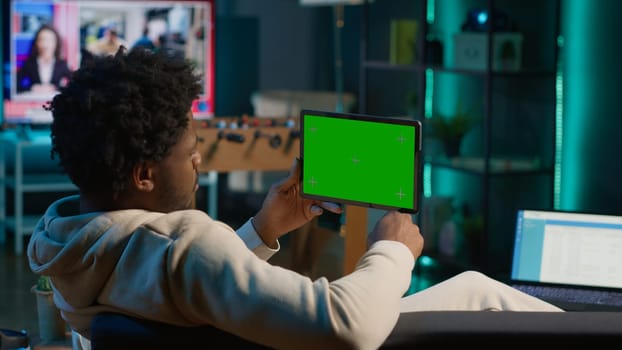 African american male freelancer holding device with mockup chroma key screen, relaxing on his couch next to neon lights and smart tv setup. Young adult feels happy at his home. Camera A.
