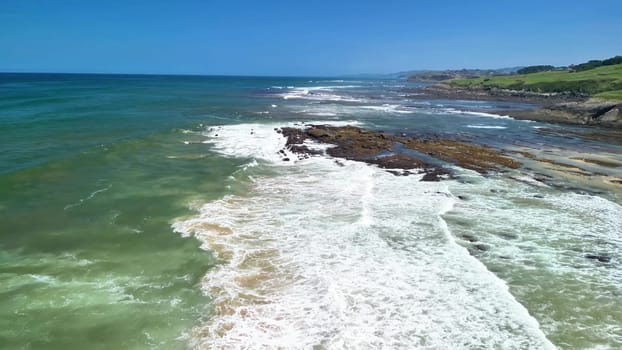 Enjoy the stunning aerial view of the coastline with waves, rocks, and greenery under a clear blue sky