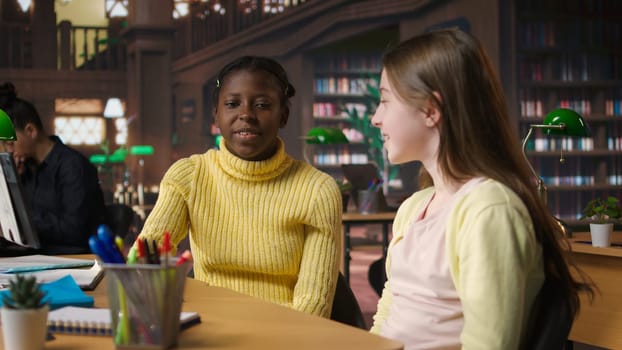 Cute preadolescent girls gossiping and hugging each other in the library, takes break from school work and enjoying assignment together. Young scholars laughing and enjoying chat. Camera A.