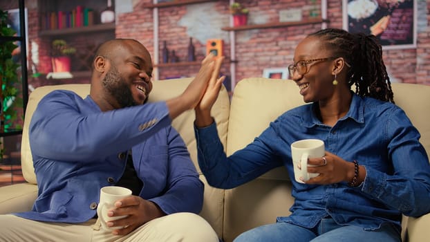 Black couple bonding on the couch unwinding with warm drinks after a long day. Smiling man and woman relaxing in a cozy setting, the beauty of companionship and peaceful relaxation. Camera A.