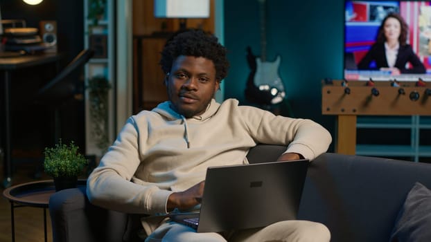 African American freelancer working remotely on a laptop under neon lights, sitting on a couch in a cozy loft apartment. Relaxed man in a stylish home highlights modern productivity. Camera A.