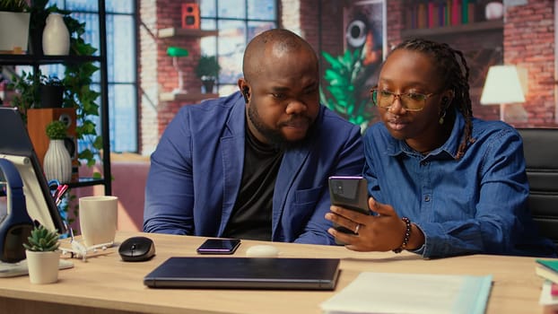 Young black couple sharing ear buds to listen to webinar on smartphone, working together on their freelancing tasks at home. Man and woman discussing about a work project. Camera A.