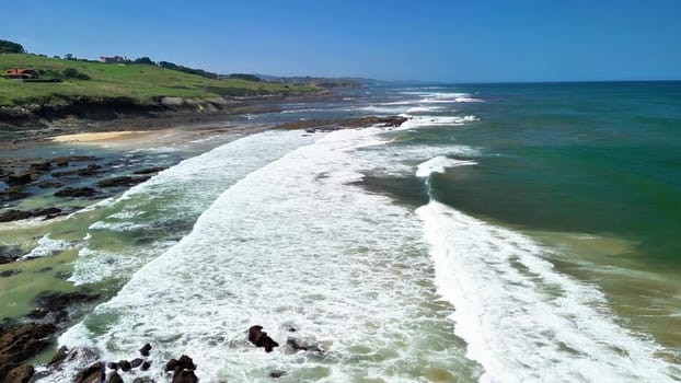 Stunning aerial view of a beautiful coastline, waves crashing against lush green hills under a clear blue sky