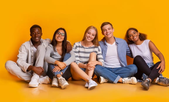 Multiracial joyful students sitting on floor, embracing and having fun over yellow studio background, students lifestyle concept, panorama