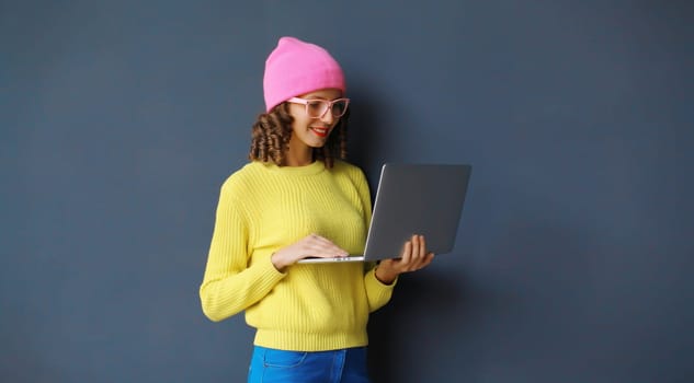 Happy modern young woman working on a laptop, freelancer girl looking at computer in colorful clothes standing against a dark wall background