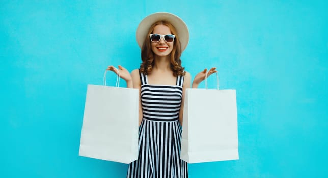 Fashion, purchase. Portrait of stylish beautiful happy young woman posing with white shopping bags, girl wear summer hat, dress standing on bright blue background