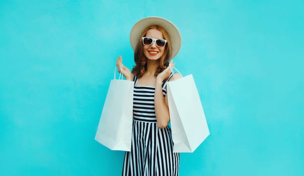 Fashion, purchase. Portrait of stylish beautiful happy young woman posing with white shopping bags, girl wear summer hat, dress standing on bright blue background
