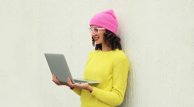 Happy modern young woman working on a laptop, freelancer girl looking at computer in colorful clothes standing against a white wall background