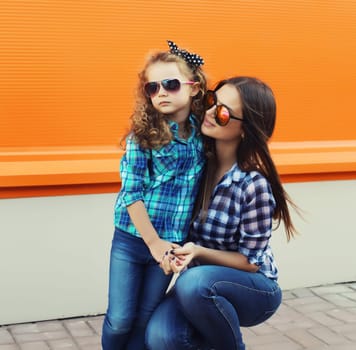 Beautiful stylish mother and child daughter together, happy mom with kid girl in glasses, casual clothing in the city on an orange background