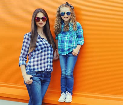 Beautiful stylish mother and child daughter together, happy mom with kid girl in glasses, casual clothing in the city on an orange background