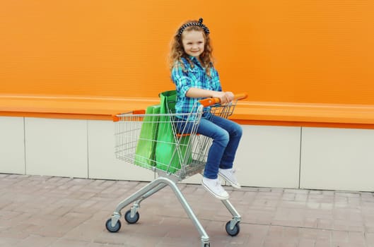 Happy little girl child sitting in trolley cart and colorful shopping bags, kid on city street
